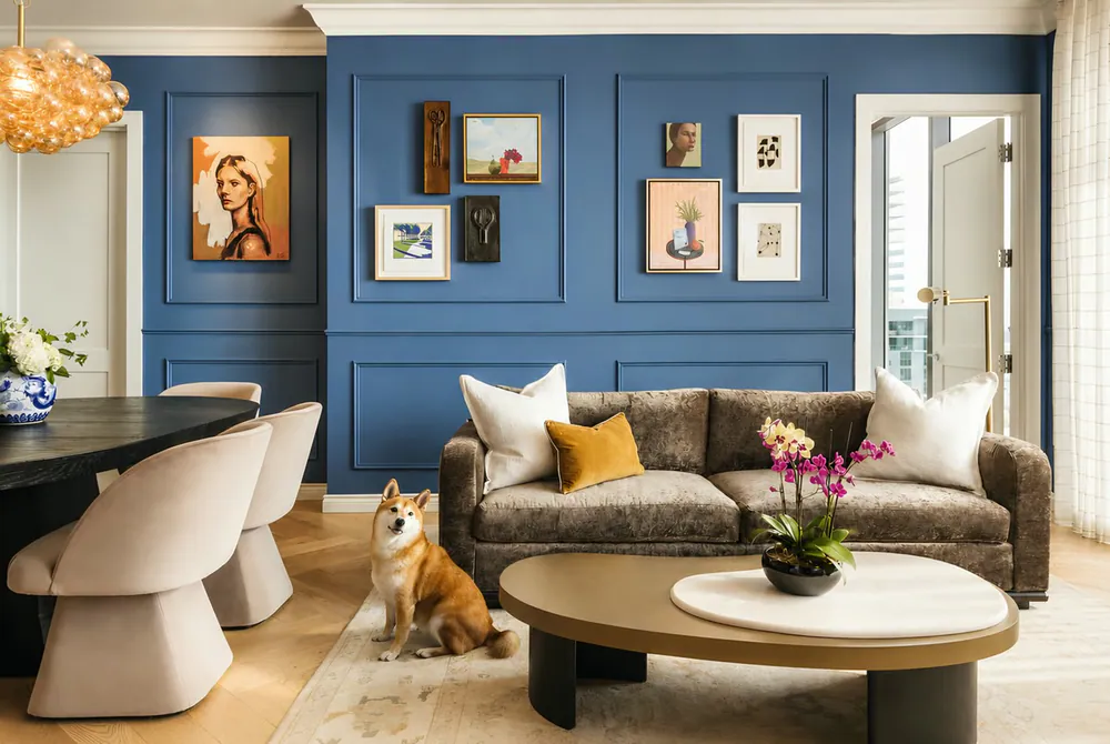 Living room with blue paneled walls, a gallery of original art, mink velvet sofa and oval bronze coffee table, at Art House in Downtown St. Petersburg. Interior design by MJ Designs