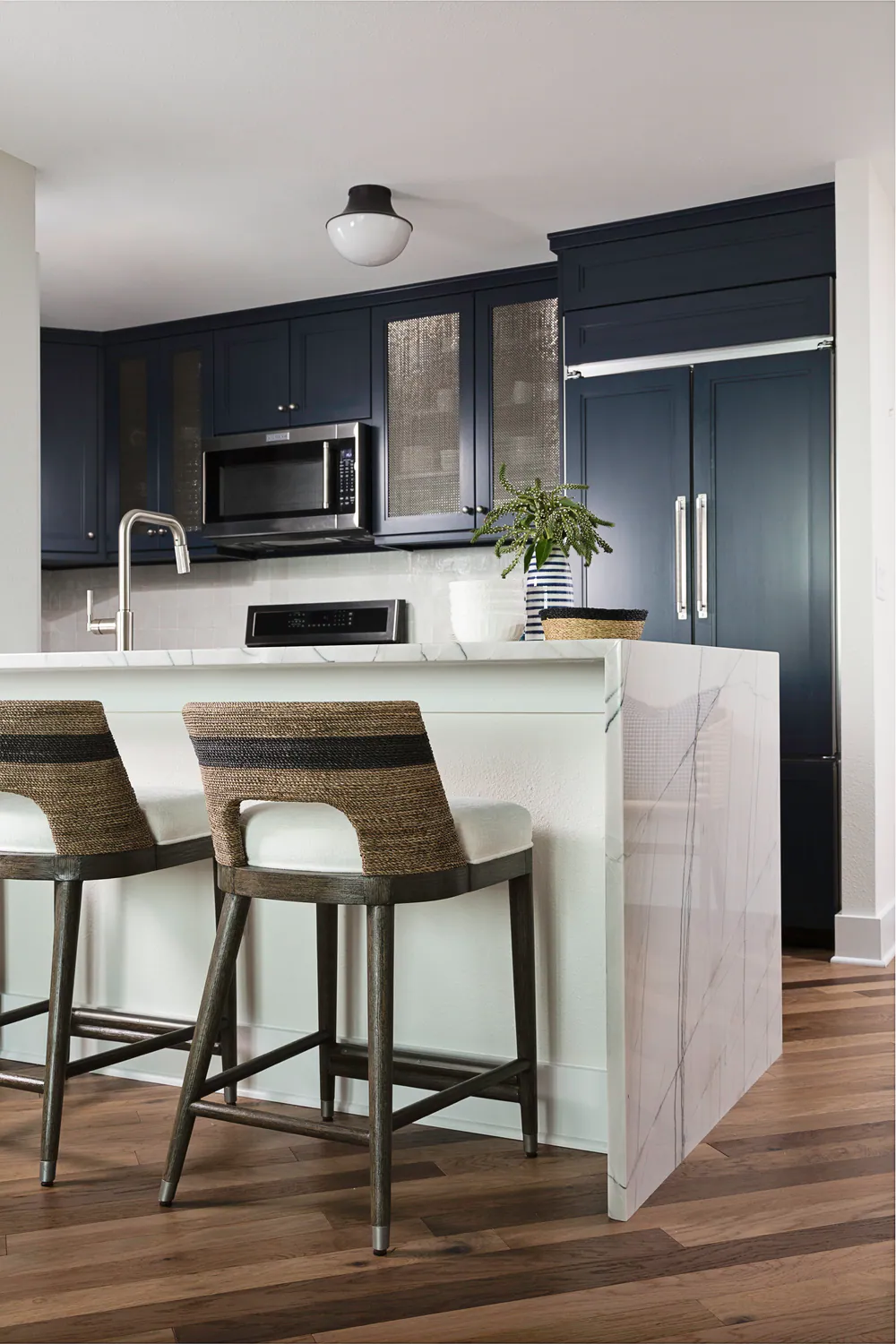 Kitchen with a veined stone waterfall peninsula, woven counter stools and navy cabinetry, at Bayshore in Tampa. Interior design by MJ Designs