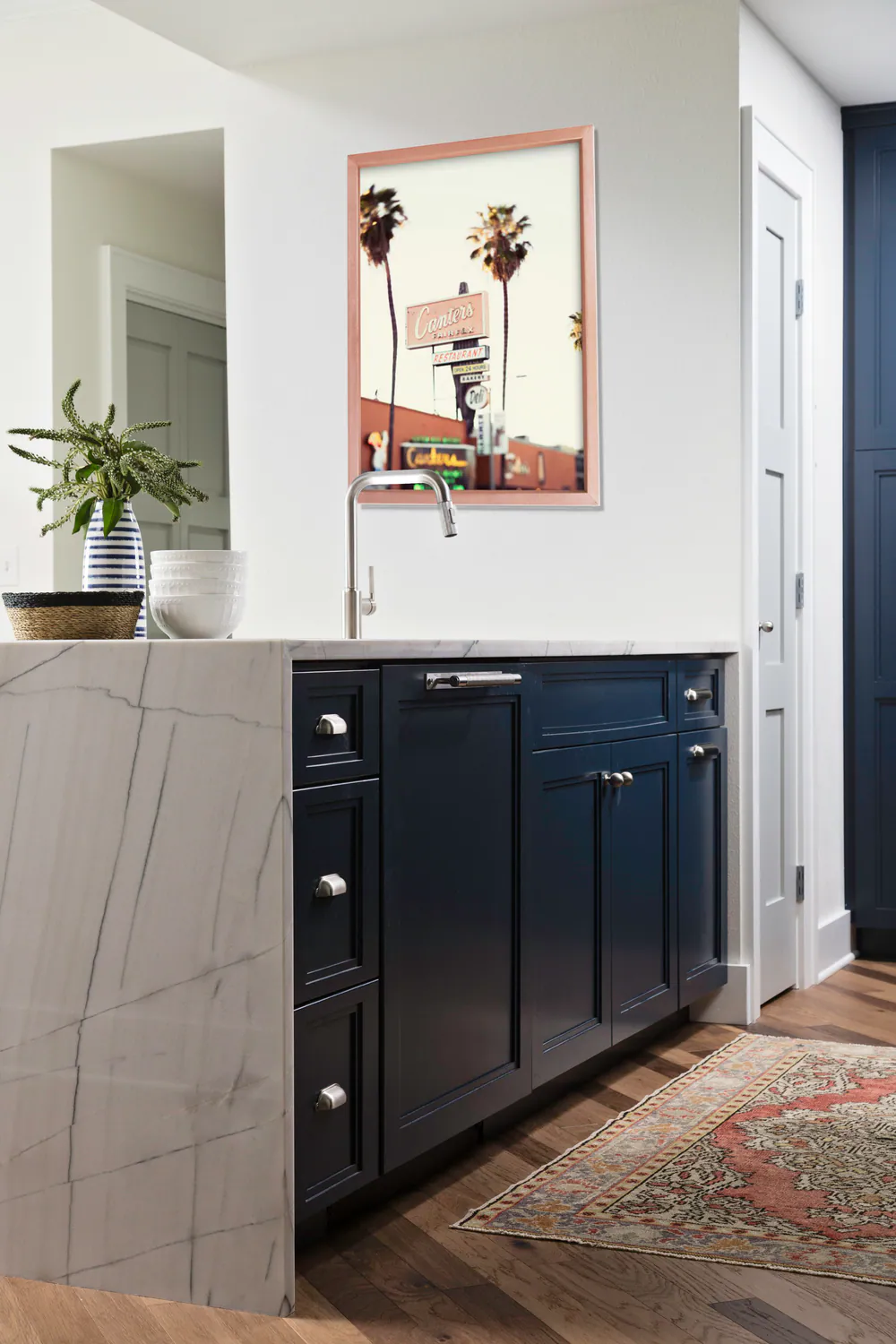 Navy base cabinets and a veined stone waterfall peninsula below a framed photograph of palms, at Bayshore in Tampa. Interior design by MJ Designs