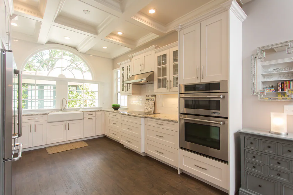 White kitchen with stainless wall ovens, glass-front cabinets, a vent hood and an arched window over the farmhouse sink, at Brightwaters in St. Petersburg. Interior design by MJ Designs