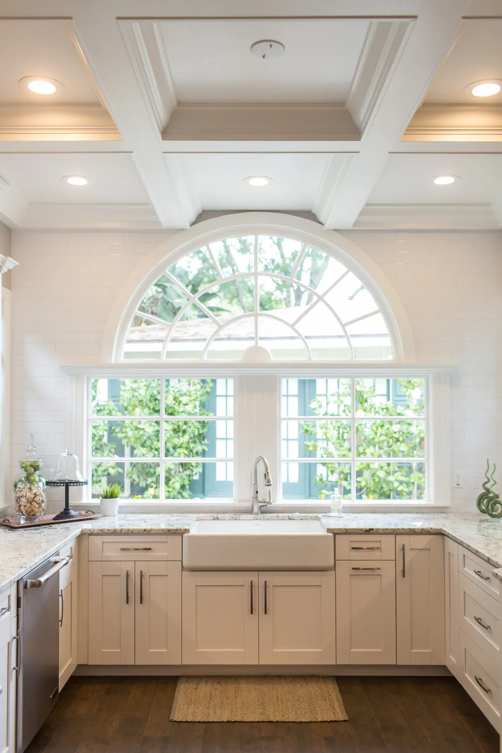 Farmhouse sink beneath an arched transom window, with white subway tile, shaker cabinetry and a coffered ceiling, at Brightwaters in St. Petersburg. Interior design by MJ Designs