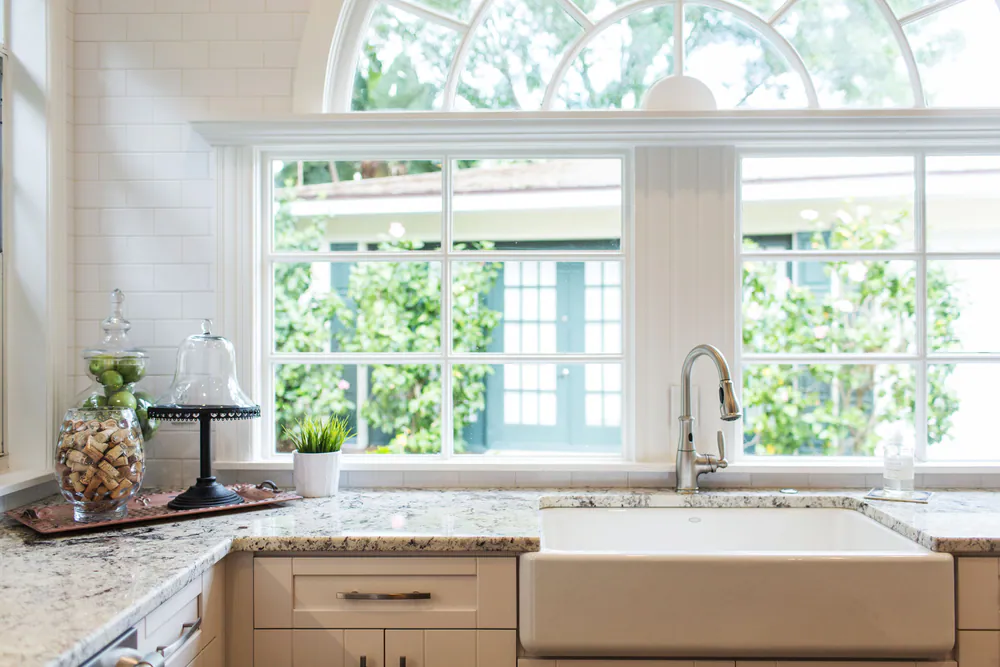 Farmhouse sink and speckled granite counters beneath divided-light windows and an arched transom, at Brightwaters in St. Petersburg. Interior design by MJ Designs