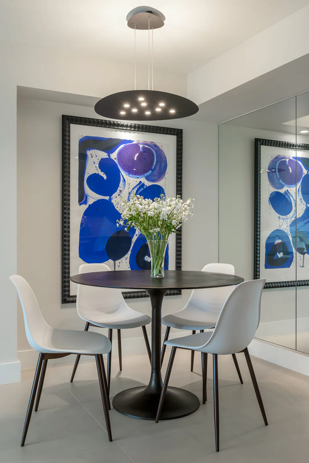 Round black pedestal table with white chairs under a disc pendant, blue abstract and mirrored wall, at Gulf Beaches Condo in Pinellas County. Interior design by MJ Designs