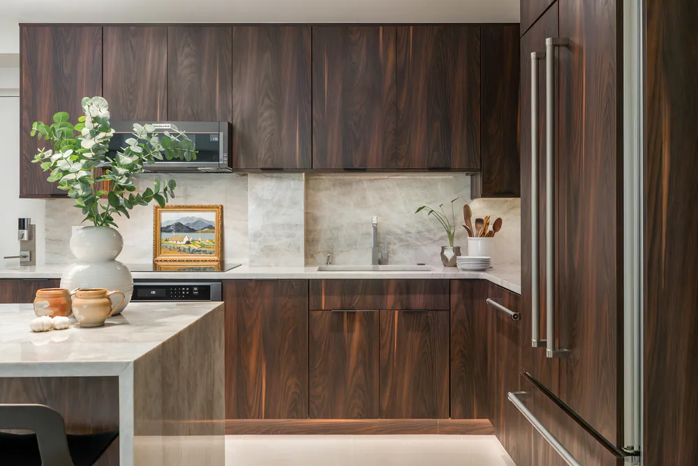 Kitchen with walnut-toned slab cabinetry, veined stone backsplash and waterfall island, at Gulf Beaches Condo in Pinellas County. Interior design by MJ Designs
