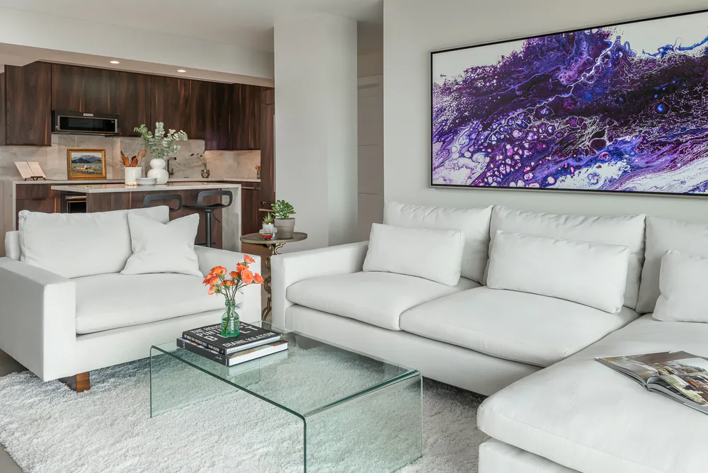 Living room with white sectional and armchair, glass coffee table and the open kitchen beyond, at Gulf Beaches Condo in Pinellas County. Interior design by MJ Designs