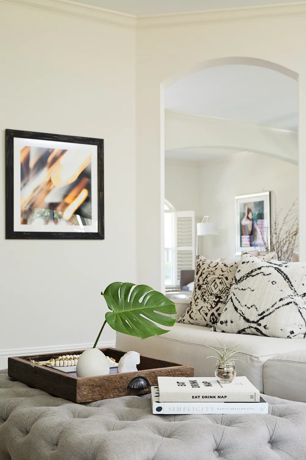 Tufted ottoman with wood tray and monstera leaf, view through an arch to the sitting room, at Hyde Park in Tampa. Interior design by MJ Designs